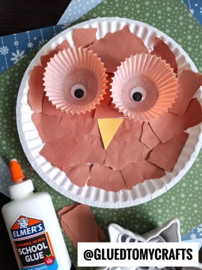 A paper plate owl craft featuring torn brown paper, cupcake liners for eyes, googly eyes, and an orange beak. Elmer’s school glue, brown paper scraps, and a white cookie cutter surround the project. Text reads @GLUEDTOMYCRAFTS.
