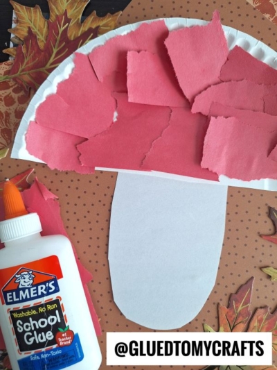 A fun FALL CRAFTS FOR KIDS project: a paper plate shaped like a red and white mushroom, decorated with torn red paper and Elmer’s glue, displayed on a brown dotted background with autumn leaves.