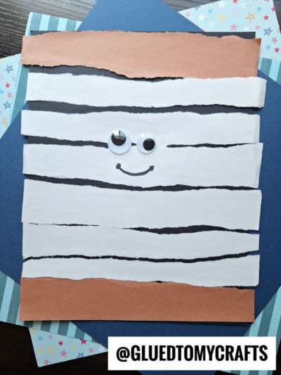 A fun mummy craft made from strips of torn white and brown paper, googly eyes, and a drawn smile. With patterned and solid backgrounds, this is one of the best summer crafts for kids to spark creativity.