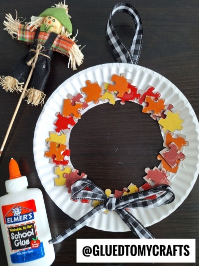 A fall-themed wreath made from a paper plate and colored puzzle pieces, decorated with a black-and-white ribbon bow. Perfect for FALL CRAFTS FOR KIDS! Nearby are Elmer’s glue and a scarecrow decoration on a stick. @GLUEDTOMYCRAFTS is written below.