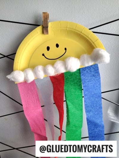 A yellow paper plate, decorated as a smiling sun with black eyes and mouth, features cotton clouds and colorful crepe paper streamers hanging below—perfect for cheerful crepe paper craft ideas. It’s clipped to a string and tagged with “@GLUEDTOMYCRAFTS.”.
