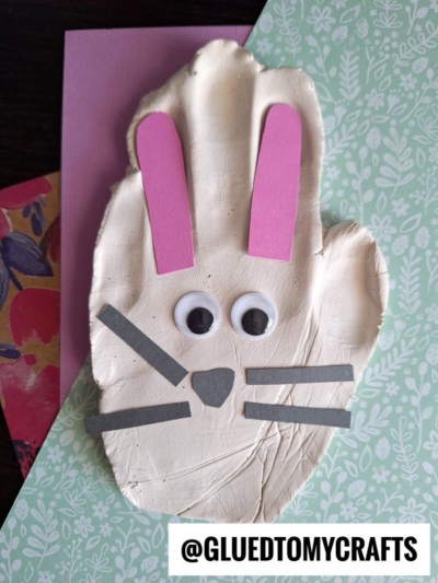 A bunny face crafted from a white handprint clay ornament features pink paper ears, googly eyes, a small gray paper nose, and gray whiskers. It rests on patterned paper with @GLUEDTOMYCRAFTS text.