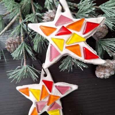 Two star-shaped ornaments decorated with colorful triangular tiles in red, orange, and yellow shades hang on a pine branch with small pinecones. Text at the bottom reads @GLUEDTOMYCRAFTS.