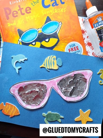 Children’s craft project inspired by Pete the Cat and His Magic Sunglasses. Features a blue paper with a cut-out of sunglasses, filled with foil, and decorated with ocean animal stickers. Elmer’s glue and book are nearby.