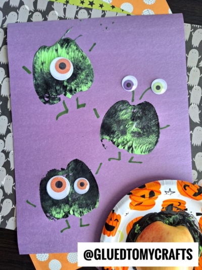 Purple paper with three green monster prints made from an apple stamped craft, each with big googly eyes and marker-drawn arms and legs. A Halloween-themed plate and orange napkin are in the background.
