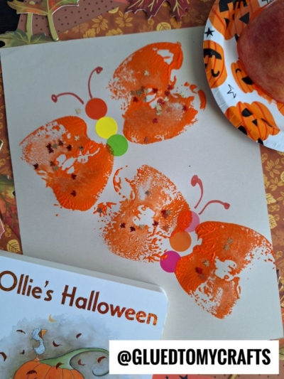 A fall-themed butterfly craft made from orange pumpkin and apple stamped craft shapes, with added antennae and colorful circles for a caterpillar on beige paper. Nearby are a Halloween-themed plate and a children’s book titled Ollie’s Halloween.