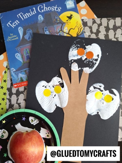 A Halloween craft scene with a Ten Timid Ghosts book, a black paper handprint ghost tree decorated with orange and yellow dots, an APPLE STAMPED CRAFT on a paper plate, and ghost-themed papers in the background.