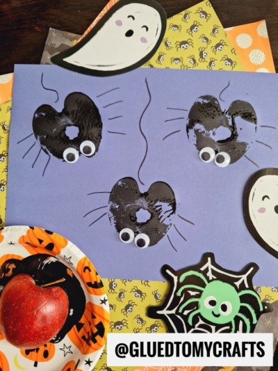 A purple paper features an apple stamped craft with three black heart-shaped mice made from apple stamps, each with googly eyes and drawn-on tails, surrounded by Halloween-themed decorations and an apple on a pumpkin plate.