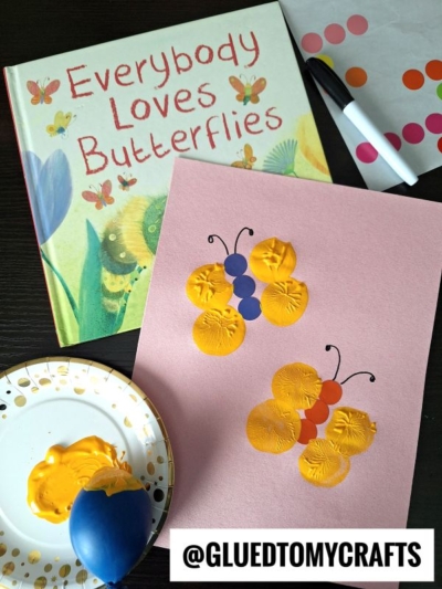 A butterfly-themed children’s book and a pink paper display insect craft ideas: butterfly crafts made with yellow paint balloon stamps and colored dot stickers. Paint, a balloon, and stickers are arranged nearby; @GLUEDTOMYCRAFTS label at the bottom.