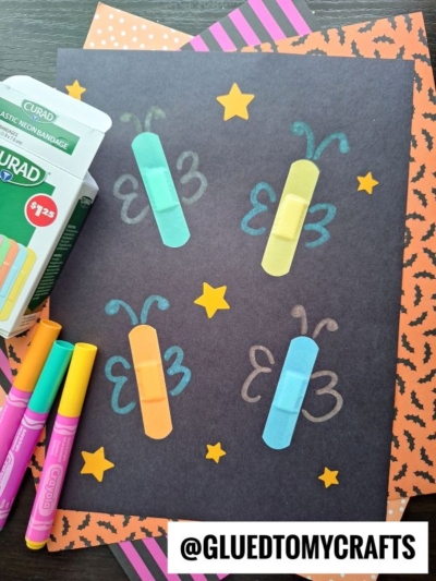 A black sheet of paper decorated with colorful bandages arranged as butterfly bodies, each with drawn wings, surrounded by yellow stars. Neon markers, patterned paper, and a box of bandages are nearby.