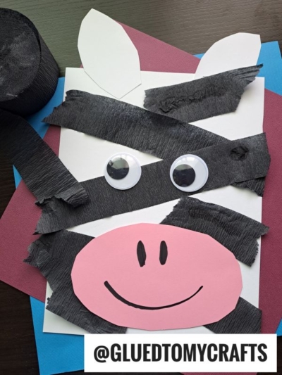 A zebra craft made from black crepe paper strips, large googly eyes, and a pink paper snout with a smile rests on a white background with colored sheets—perfect for anyone looking for fun crepe paper craft ideas. Crepe paper roll and handle @GLUEDTOMYCRAFTS visible.
