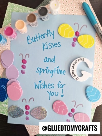 A light blue paper decorated with foam butterflies and eggs, paint pots, a pen, and a ceramic letter G. Handwritten text reads: Butterfly kisses and springtime wishes for you! @GLUEDTOMYCRAFTS.