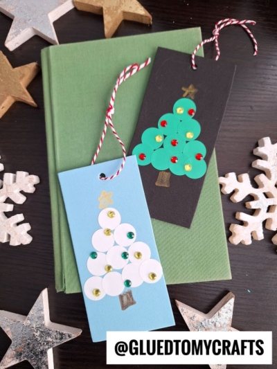 Two handmade Christmas gift tags, one blue and one black, each decorated with a paper tree and sequins, rest on a green book. Wooden stars and snowflake ornaments are scattered around—perfect inspiration for coding label crafts.