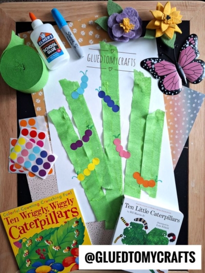 A flat lay of childrens caterpillar craft materials: green crepe paper, construction paper, glue, scissors, colorful dot stickers, foam flowers, a butterfly, coding label crafts examples, and two caterpillar-themed books. @GLUEDTOMYCRAFTS is displayed.