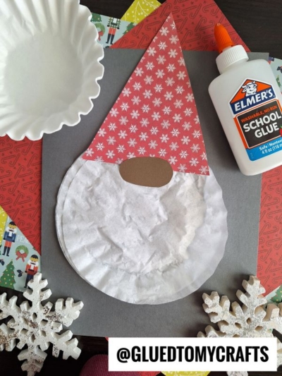 A festive Christmas gnome craft, perfect for coffee filter crafts for kids, features a red patterned hat, a coffee filter beard, brown nose, and gray paper background with glue, white snowflakes, and craft supplies scattered around.