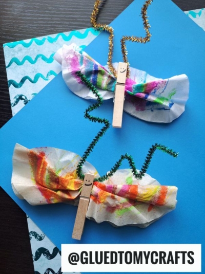 Two colorful butterfly crafts, perfect for INSECT CRAFT IDEAS, made from painted coffee filters, clothespins with smiley faces, and twisted pipe cleaners as antennae, are displayed on blue paper with a wave pattern. @GLUEDTOMYCRAFTS is written at the bottom.