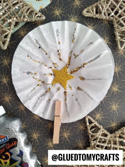 A coffee filter decorated with glitter and a gold star in the center, attached to a clothespin. Glittery star ornaments and a bottle of glitter glue surround it on a black surface with gold dots. @GLUEDTOMYCRAFTS is displayed at the bottom.