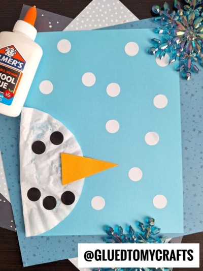 A snowman craft made from a blue paper background, white circles for snow, black dots for eyes and mouth, an orange triangle nose, a coffee filter for the face, glue bottle, and snowflake decorations—perfect coffee filter crafts for kids.