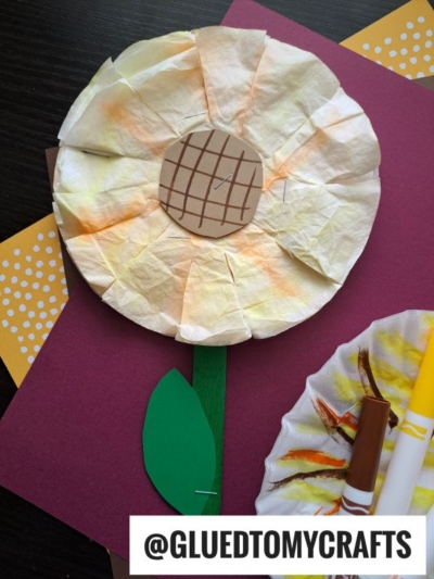 A paper plate flower craft with a brown paper center, green stem and leaf, and yellow-tinted petals, lying on maroon and yellow polka dot paper. Brown and yellow markers are nearby. @GLUEDTOMYCRAFTS is at the bottom.