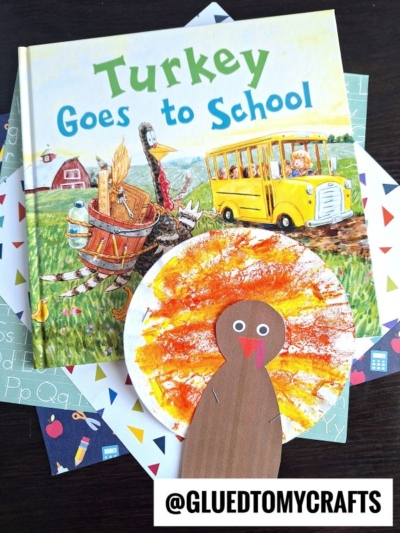 Children’s book “Turkey Goes to School” and several colorful papers are on a table, with a handmade turkey craft featuring a brown body and orange, yellow, and red painted feathers in front. @GLUEDTOMYCRAFTS is at the bottom.