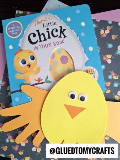 A children’s book titled “There’s a Little Chick in Your Book” is displayed with a yellow chick craft made of paper, featuring googly eyes, an orange beak, and handprint wings. @GLUEDTOMYCRAFTS is written at the bottom.