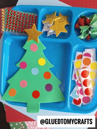 A blue tray holds a green paper Christmas tree decorated with colorful dot stickers, coding label crafts, glittery gold stars, gem stickers, paper bows, and sheets of round stickers. The Instagram handle @GLUEDTOMYCRAFTS is visible.