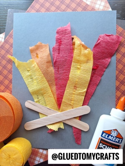 A craft project resembling a campfire made from red, orange, and yellow crepe paper strips, two craft sticks arranged like logs, and a bottle of Elmer’s glue, with patterned paper in the background.
