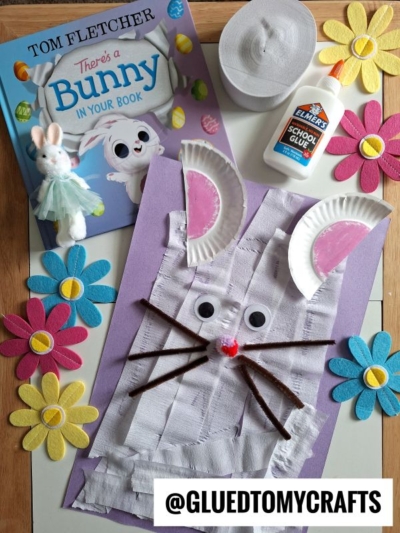 A children’s book titled “There’s a Bunny in Your Book” sits next to a bunny craft made from paper plates, googly eyes, and pipe cleaners, surrounded by colorful felt flowers and a bottle of Elmer’s glue.