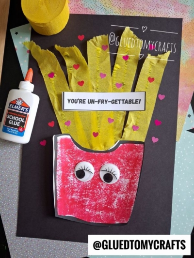 A handmade craft of crepe paper fries with googly eyes and a sign that says YOU'RE UN-FRY-GETTABLE! is shown. There is also a bottle of Elmer's school glue and a roll of yellow crepe paper nearby.