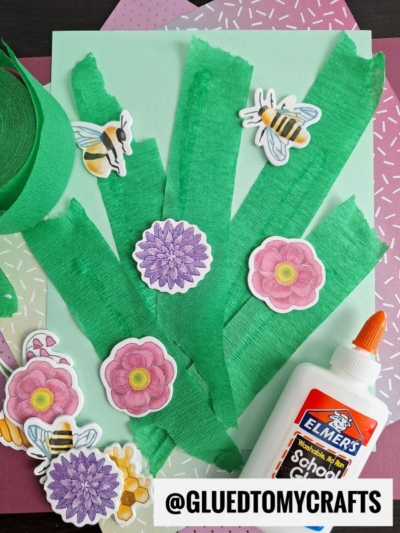 A craft project featuring green crepe paper strips as stems, flower and bee stickers, a bottle of Elmer’s glue, and colorful patterned paper in the background—perfect for anyone seeking crepe paper craft ideas. Text: @GLUEDTOMYCRAFTS at the bottom.