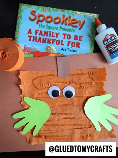 Children’s craft supplies and book: “Spookley the Square Pumpkin,” brown paper, orange crepe paper, green cut-out handprints, large googly eyes, Elmer’s glue, and fun crepe paper craft ideas. @GLUEDTOMYCRAFTS watermark.