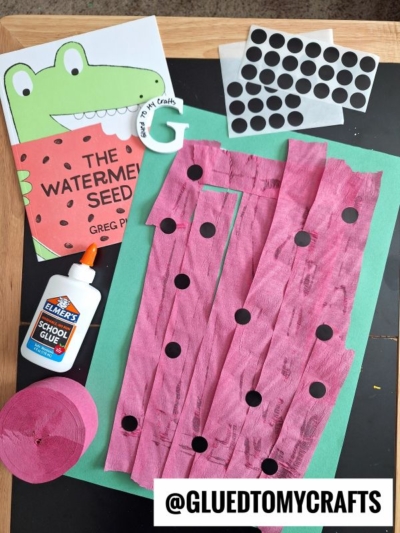 Children’s watermelon craft supplies on a table: a book with a frog on the cover, green paper, pink crepe paper strips with black dot stickers, a sheet of dot stickers, school glue, and @GLUEDTOMYCRAFTS watermark.