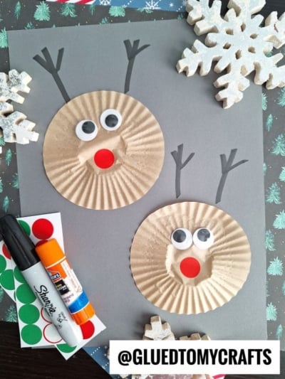 Two reindeer faces made from brown cupcake liners, googly eyes, and red dot noses are on gray paper. Surrounded by snowflake decorations, a glue stick, marker, and stickers—perfect for fun cupcake liner card crafts! @GLUEDTOMYCRAFTS at the bottom.