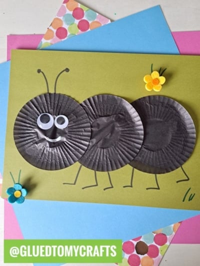 A craft ant made from three black mini paper plates on a green background, with googly eyes, drawn legs and antennae, felt flowers, colorful paper underneath—perfect inspiration for cupcake liner card crafts.