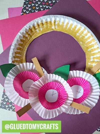 A paper plate wreath decorated with pink and white cupcake liners as flowers, green paper leaves, and brown paper strips—a fun twist on cupcake liner card crafts. Displayed on colorful backgrounds. @GLUEDTOMYCRAFTS is written at the bottom.