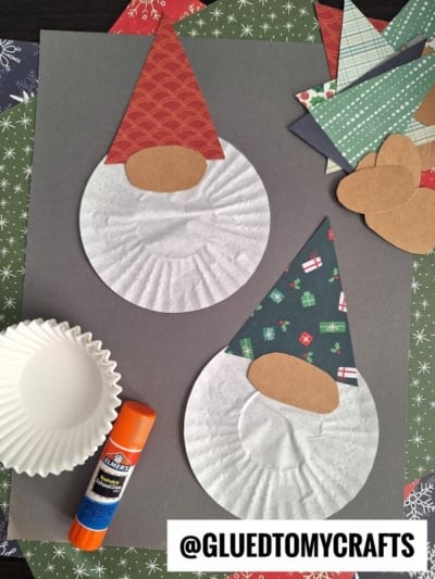 Two paper gnome crafts made with cupcake liners for bodies, patterned paper hats, and oval noses are on a dark background—perfect inspiration for cupcake liner card crafts. Nearby are more paper pieces, a glue stick, and @GLUETOMYCRAFTS.