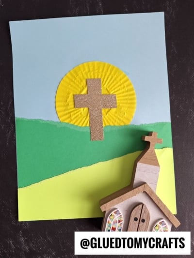 A handmade cupcake liner card craft features a brown cross on a yellow circle, set against green hills and a blue sky. A small wooden church sits in front on a dark surface. Text reads “@GLUEDTOMYCRAFTS.”.