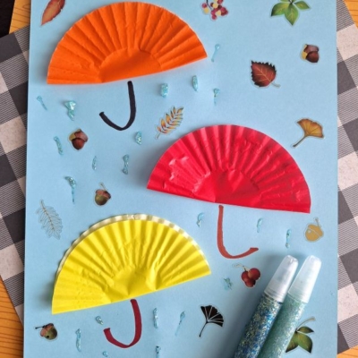 A blue paper showcases three half-circle cupcake liners in orange, red, and yellow as umbrellas—perfect for FALL CRAFTS FOR KIDS—with drawn handles, glitter raindrops, leaf stickers, and two glitter glue tubes beside it. @GLUEDTOMYCRAFTS is at the bottom.