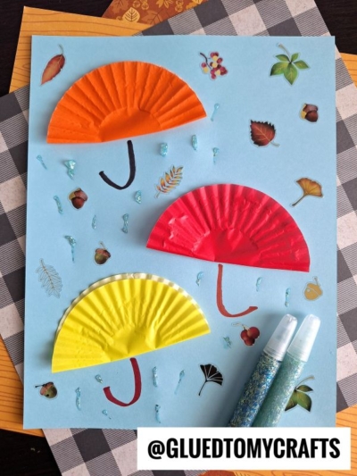 A blue paper showcases three half-circle cupcake liners in orange, red, and yellow as umbrellas—perfect for FALL CRAFTS FOR KIDS—with drawn handles, glitter raindrops, leaf stickers, and two glitter glue tubes beside it. @GLUEDTOMYCRAFTS is at the bottom.