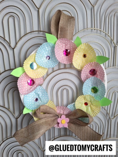 A handmade wreath decorated with pastel cupcake liners shaped like flowers, colorful gem centers, green paper leaves, and a brown ribbon bow on a textured gray background. A @GLUEDTOMYCRAFTS tag is at the bottom.