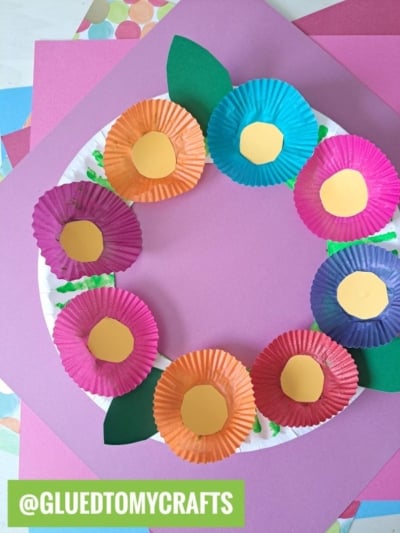 A paper plate wreath decorated with colorful cupcake liners as flowers, each with a yellow center and green paper leaves, displayed on a pink background. A green label reads @GLUEDTOMYCRAFTS.