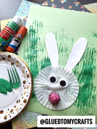 A bunny cupcake liner card craft features googly eyes, a pink pom-pom nose, and paper ears on green painted paper. Glue sticks, markers, and a plate with green paint and a fork surround it. @GLUEDTOMYCRAFTS appears at the bottom.
