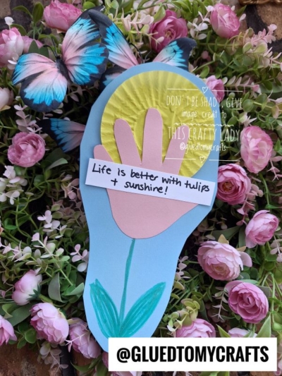 A colorful paper craft shaped like a footprint with a tulip and sun, surrounded by pink flowers and a blue butterfly. A note reads, Life is better with tulips & sunshine! and @GLUEDTOMYCRAFTS is at the bottom.