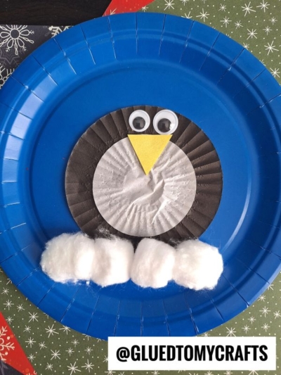 A penguin craft made from a blue paper plate, a black cupcake liner, googly eyes, yellow paper beak, and cotton balls for snow, with @GLUEDTOMYCRAFTS label at the bottom.