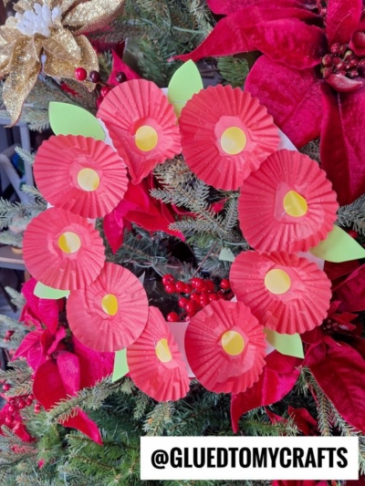 A cupcake liner flower wreath made of red liners with yellow centers and green paper leaves, displayed on a Christmas tree adorned with red poinsettias. Text @GLUEDTOMYCRAFTS appears at the bottom.