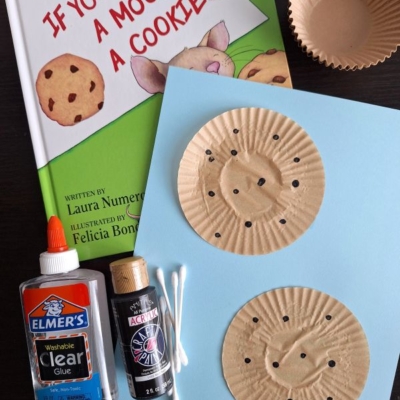 A copy of If You Give a Mouse a Cookie, glue, black paint, Q-tips, cupcake liners, and blue paper are arranged on a table, perfect for a back to school story time and craft activity.