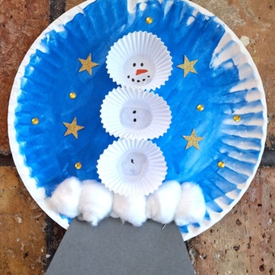 A snowman made from cupcake liners is glued to a blue painted paper plate with gold stars and gems. Cotton balls form snow at the base, and a gray paper stand completes the snow globe craft. @GluedToMyCrafts is written below.