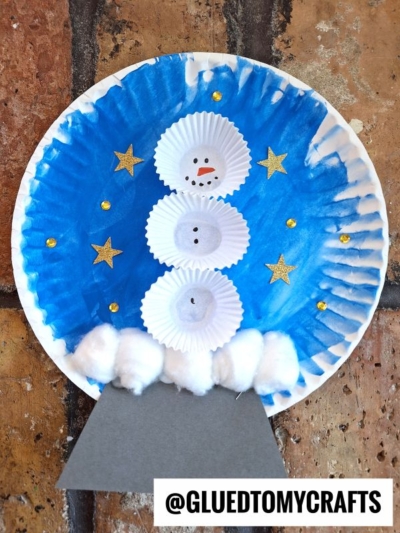 A snowman made from cupcake liners is glued to a blue painted paper plate with gold stars and gems. Cotton balls form snow at the base, and a gray paper stand completes the snow globe craft. @GluedToMyCrafts is written below.