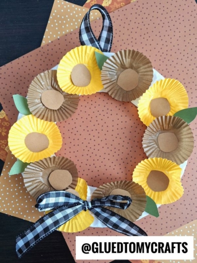 A cupcake liner flower wreath made from yellow and brown paper liners, green leaves, and a black plaid ribbon. Arranged like flowers, the wreath sits on patterned orange paper. @GLUEDTOMYCRAFTS is written at the bottom.