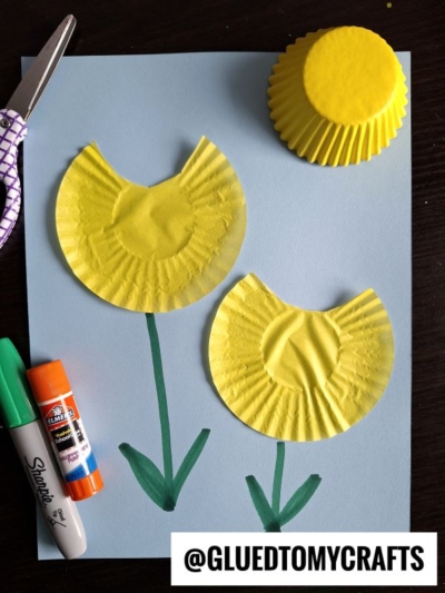 A blue paper displays two yellow cupcake liners cut in half to resemble flowers with green stems and leaves; a marker, glue stick, scissors, and an unused yellow liner are nearby. @GLUEDTOMYCRAFTS is written at the bottom.
